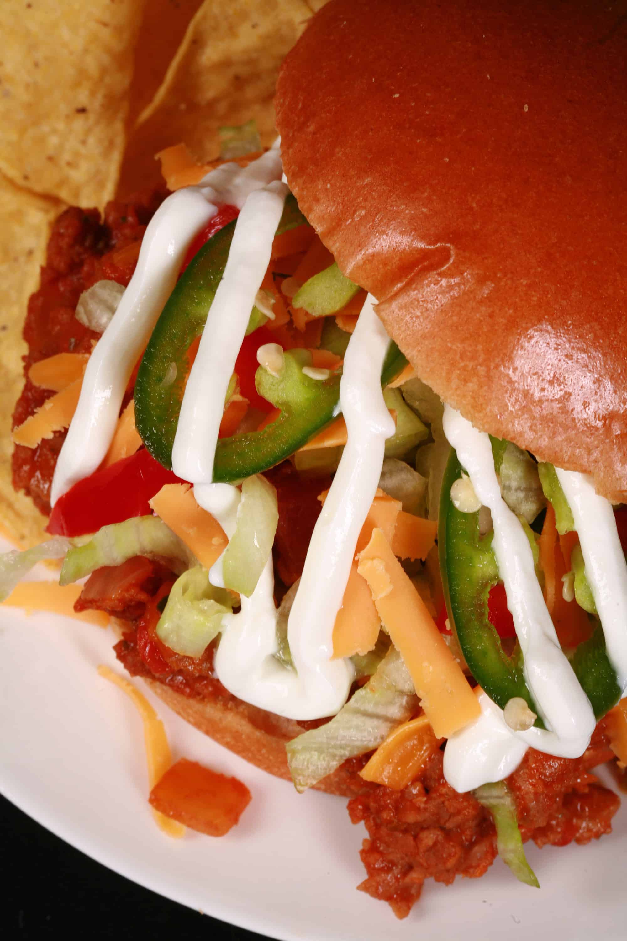 A Taco sloppy joes sandwich, with taco meat, lettuce, cheese, salsa, sour cream, and jalapeno slices, on a plate with tortilla chips.