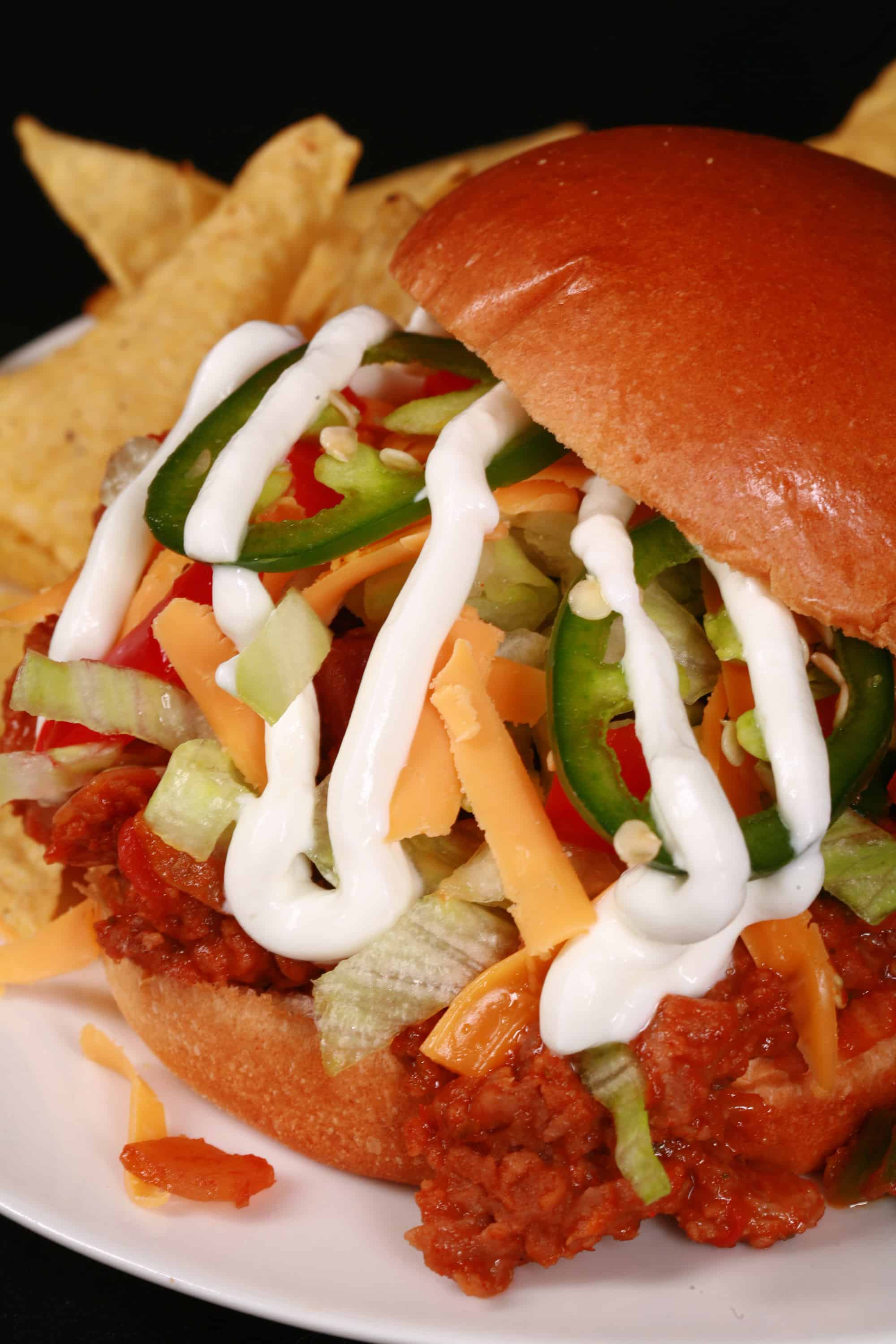 A Taco themed sloppy joe sandwich, with taco meat, lettuce, cheese, salsa, sour cream, and jalapeno slices, on a plate with tortilla chips.