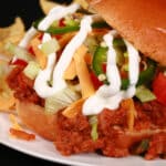 A Taco sloppy joes sandwich, with taco meat, lettuce, cheese, salsa, sour cream, and jalapeno slices, on a plate with tortilla chips.