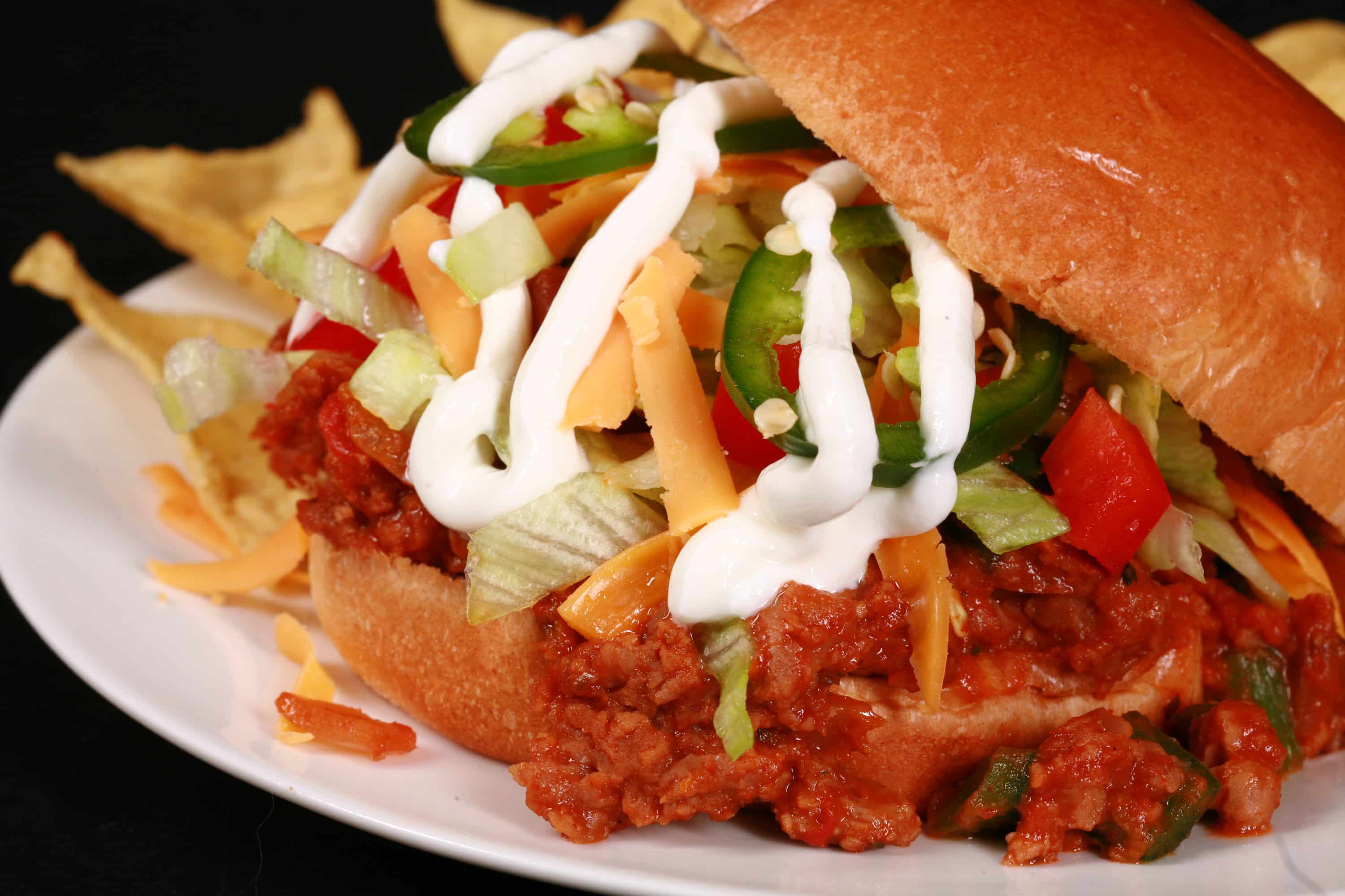 A Taco sloppy joes sandwich, with taco meat, lettuce, cheese, salsa, sour cream, and jalapeno slices, on a plate with tortilla chips.