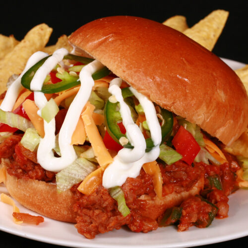 A Taco themed sloppy joe sandwich, with taco meat, lettuce, cheese, salsa, sour cream, and jalapeno slices, on a plate with tortilla chips.