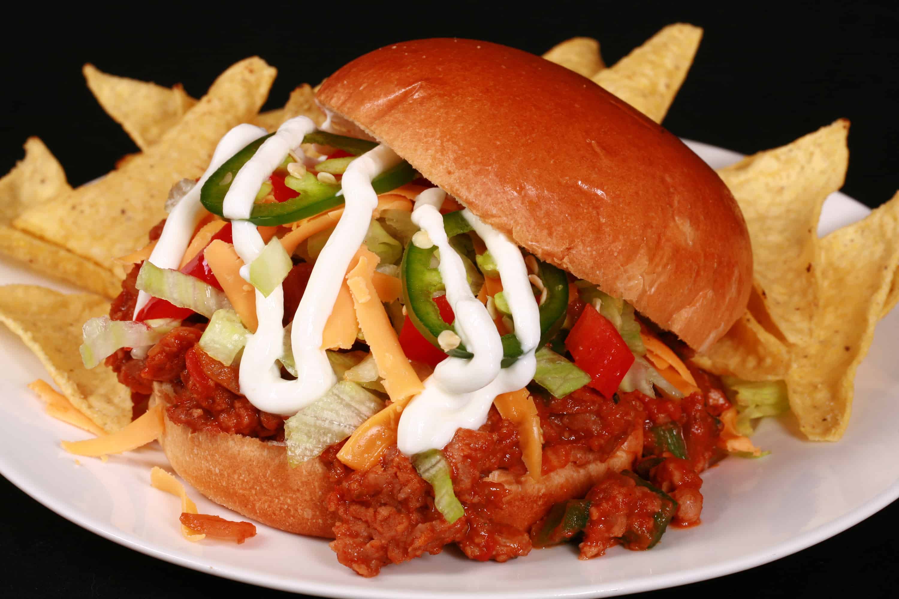 A Taco themed sloppy joe sandwich, with taco meat, lettuce, cheese, salsa, sour cream, and jalapeno slices, on a plate with tortilla chips.