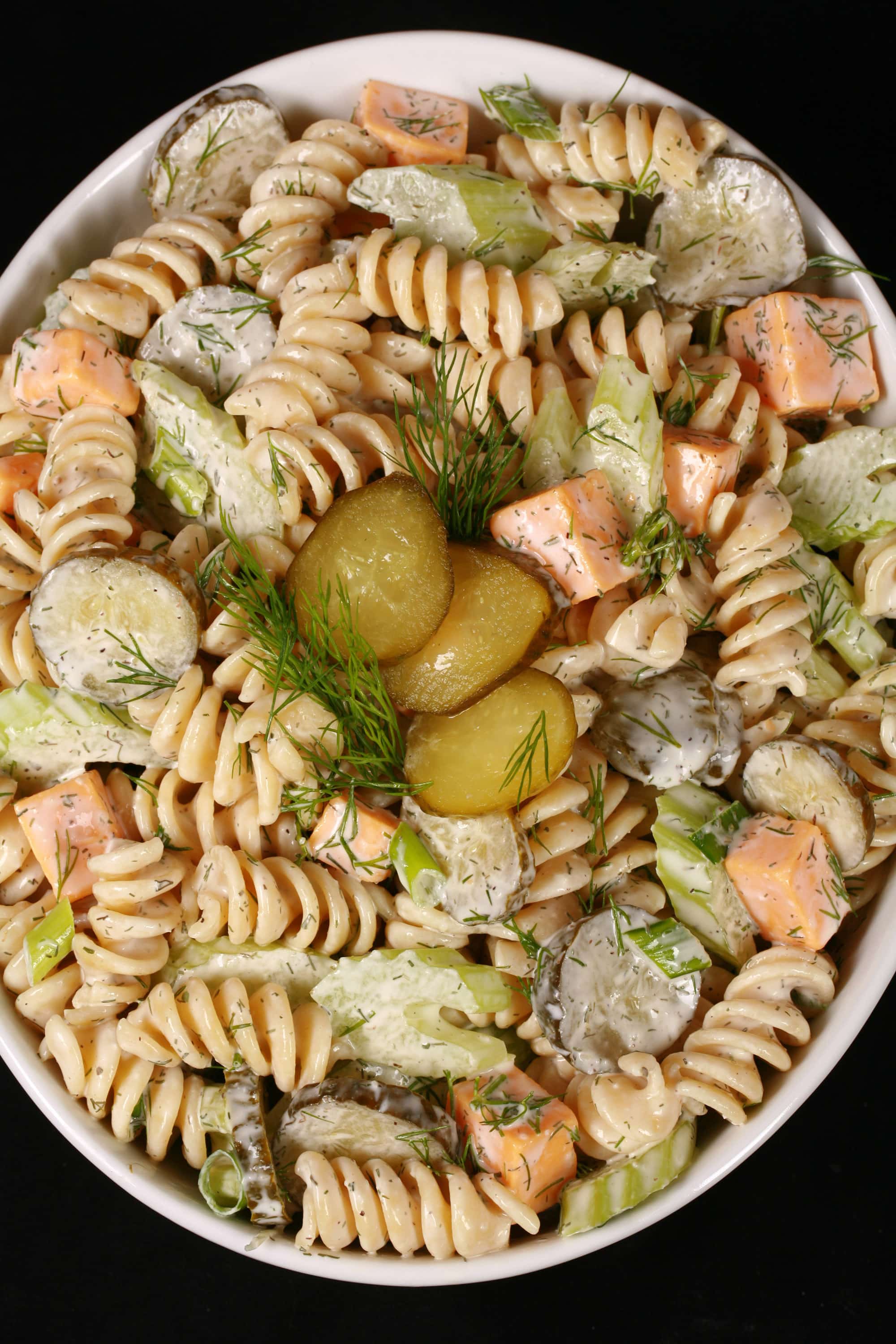 A bowl of dill pickle pasta salad, with slices of pickles, green onion, and celery, fresh dill, and cubes of cheese in a creamy sauce.