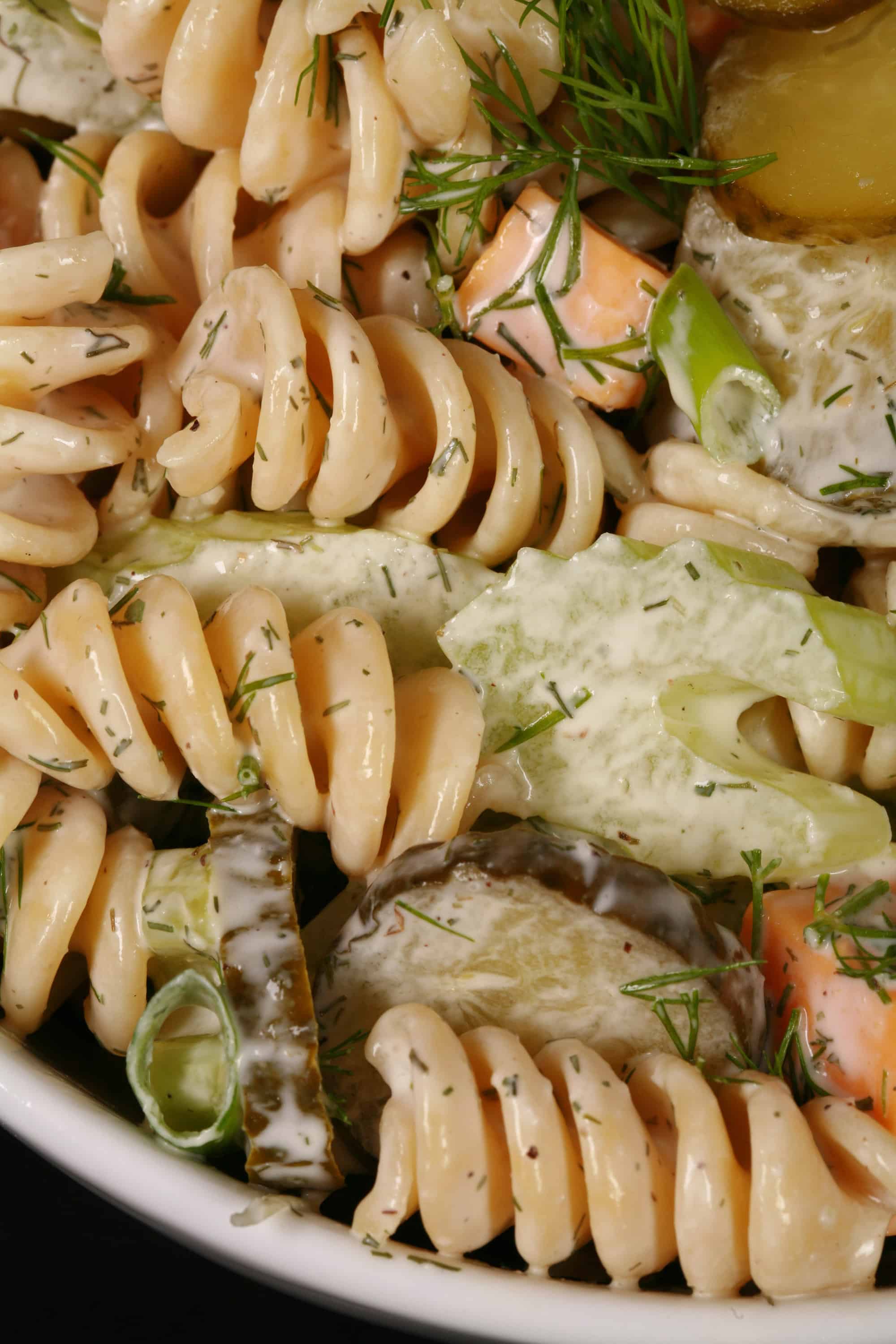 A bowl of dill pickle macaroni salad, with slices of pickles, green onion, and celery, fresh dill, and cubes of cheese in a creamy sauce.