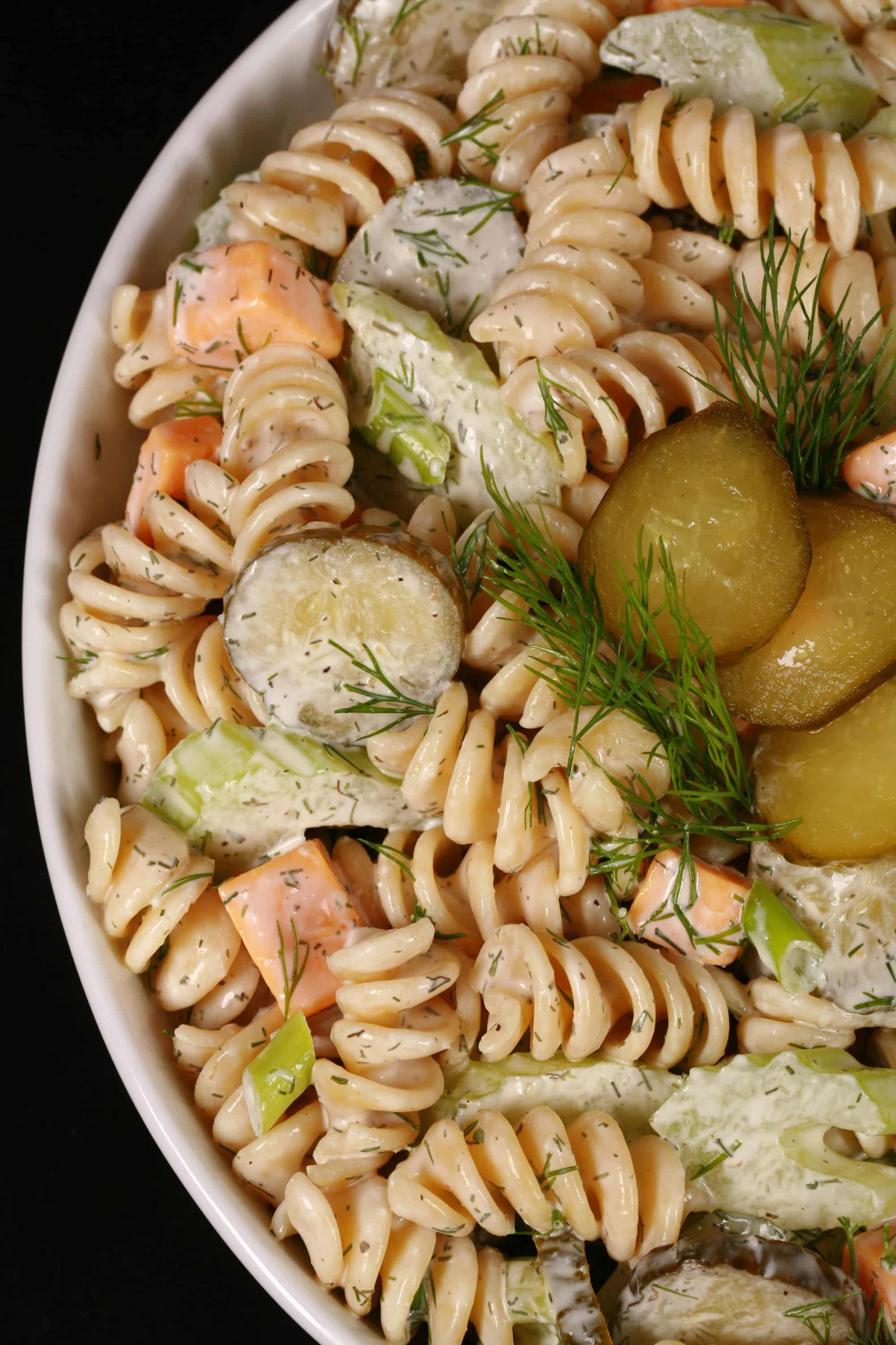 A bowl of dill pickle macaroni salad, with slices of pickles, green onion, and celery, fresh dill, and cubes of cheese in a creamy sauce.