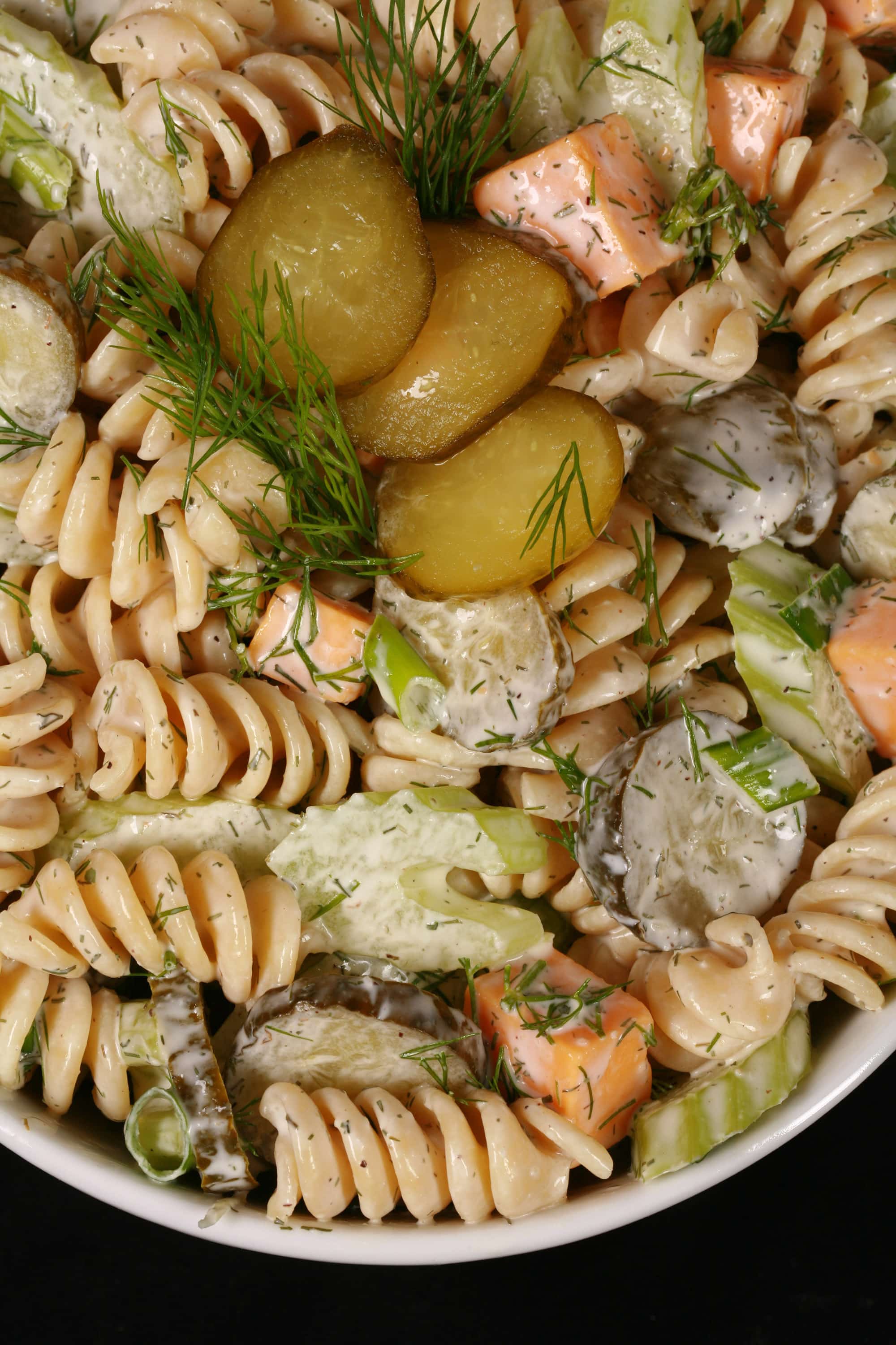 A bowl of dill pickle pasta salad, with slices of pickles, green onion, and celery, fresh dill, and cubes of cheese in a creamy sauce.