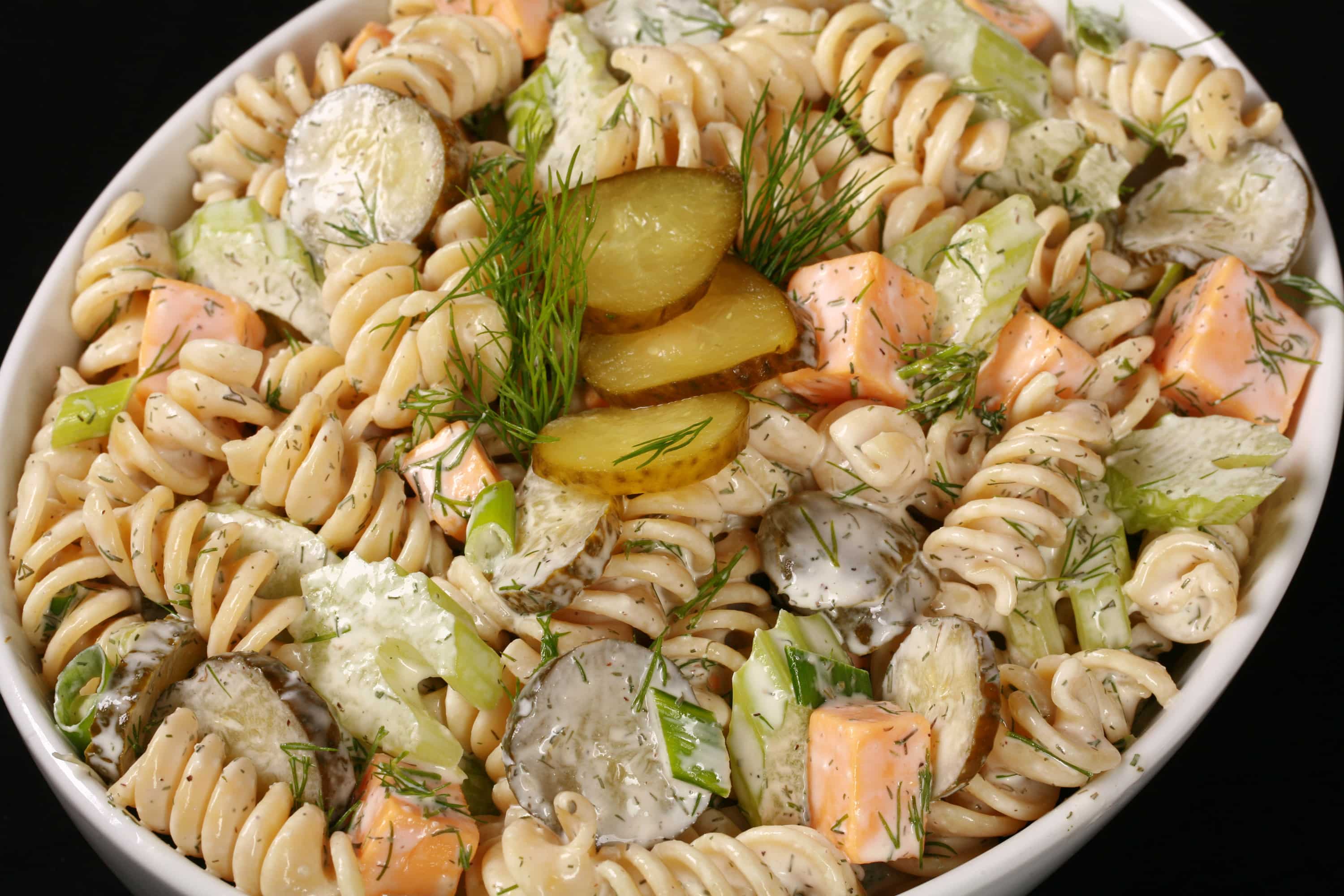 A bowl of dill pickle macaroni salad, with slices of pickles, green onion, and celery, fresh dill, and cubes of cheese in a creamy sauce.