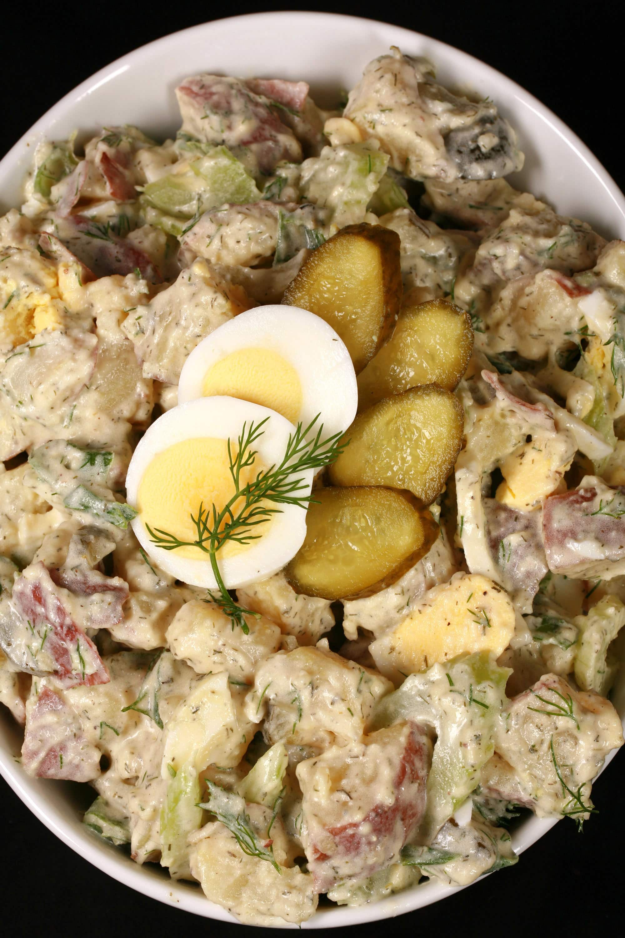 A serving bowl of potato salad with pickles, boiled eggs, celery, and dill.