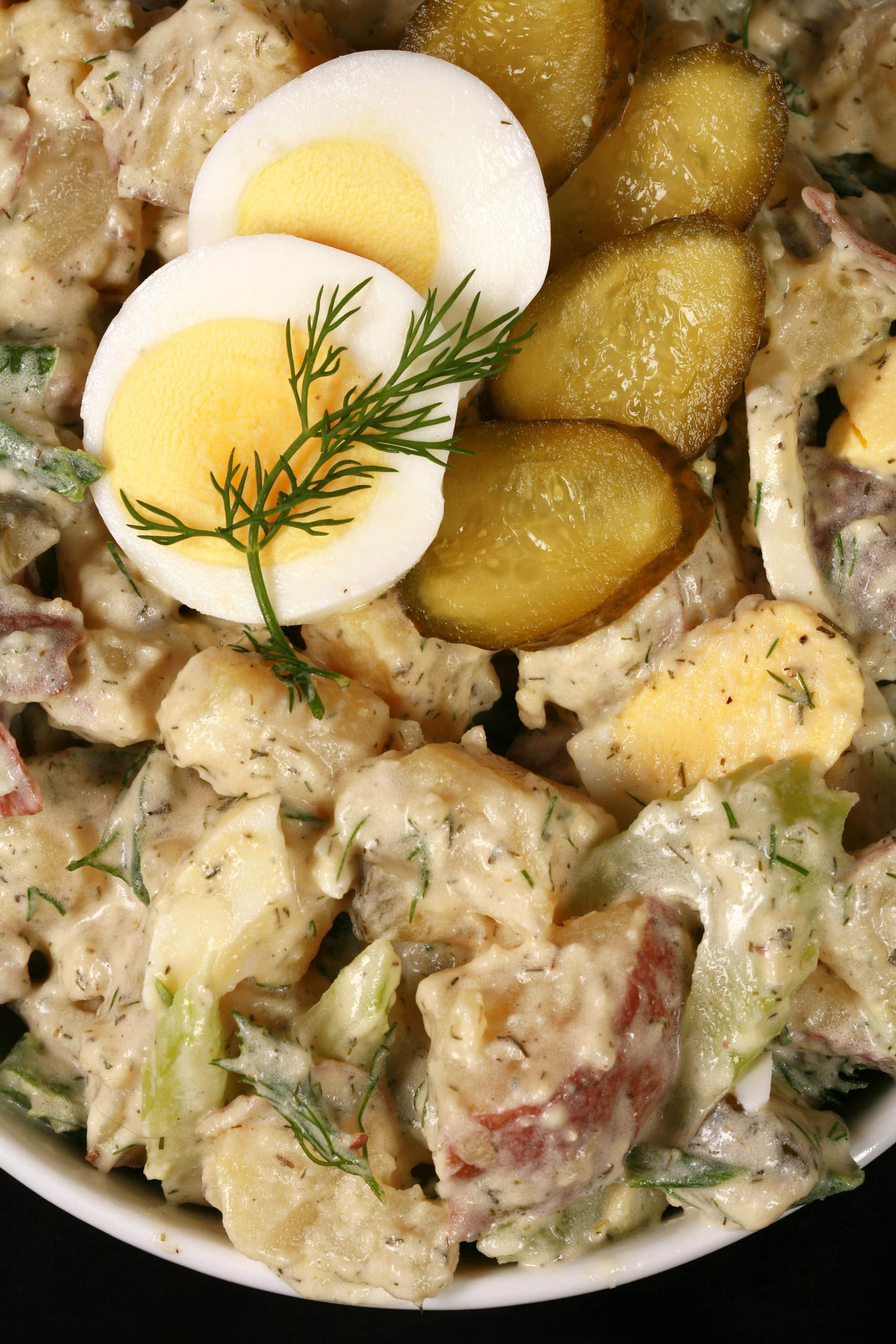 A serving bowl of potato salad with pickles, boiled eggs, celery, and dill.