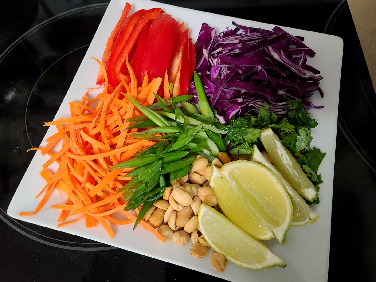 A plate with red pepper, carrot, green onions, cabbage, limes, and peanuts.