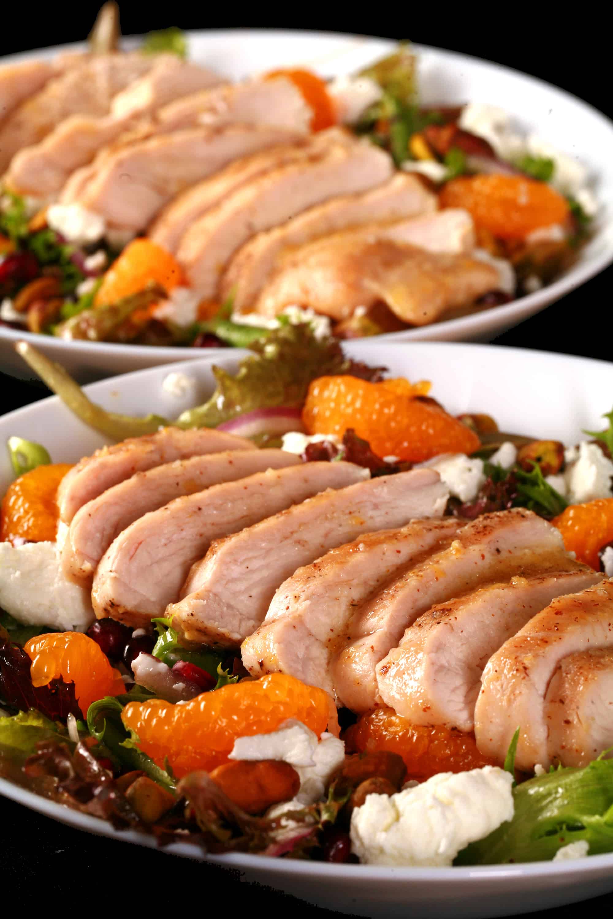 2 bowls of orange glazed chicken salad with mandarin orange segments, pomegranates, goat cheese, and pistachios.