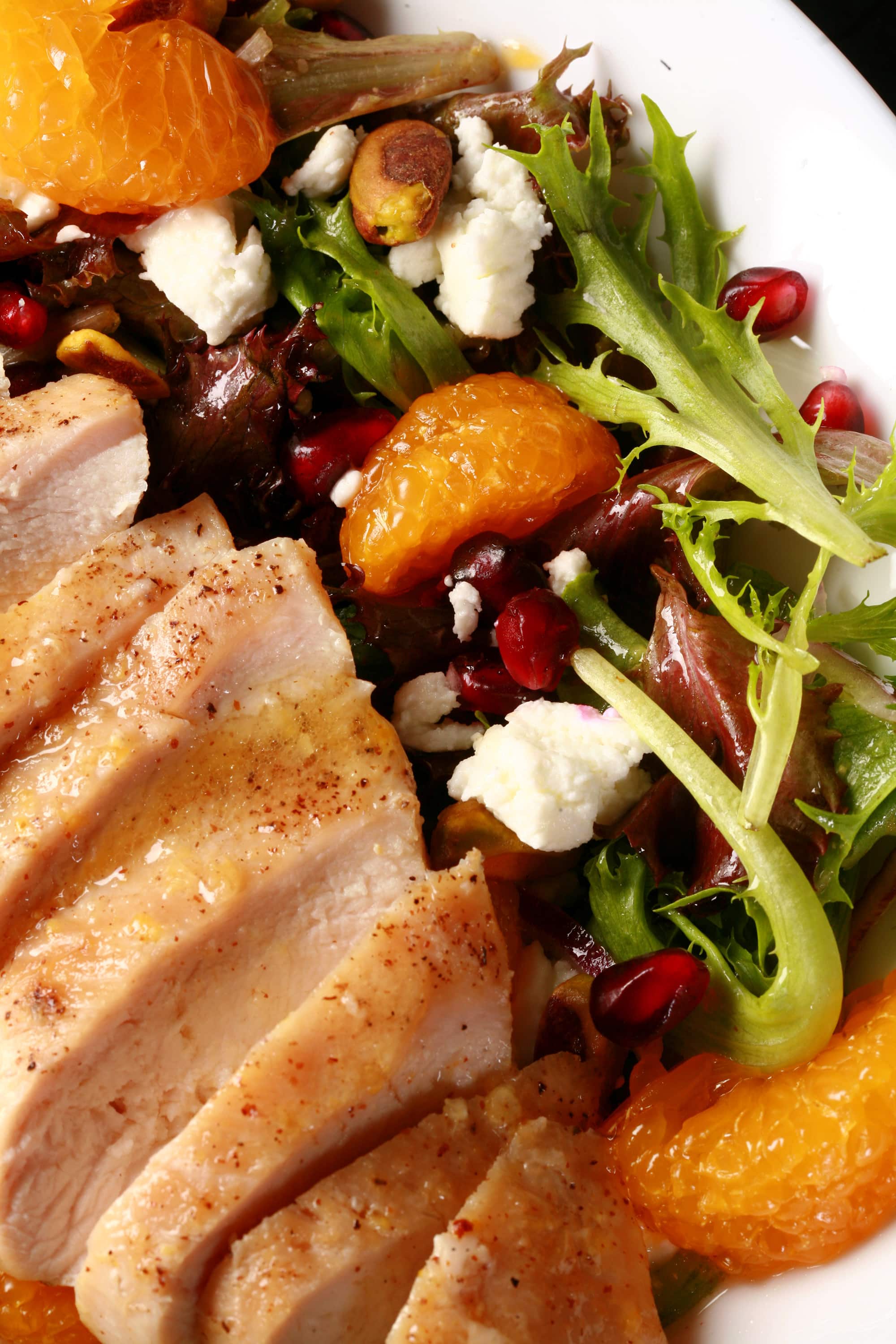 A bowl of Christmas salad with orange glazed chicken, mandarin orange segments, pomegranates, goat cheese, and pistachios.