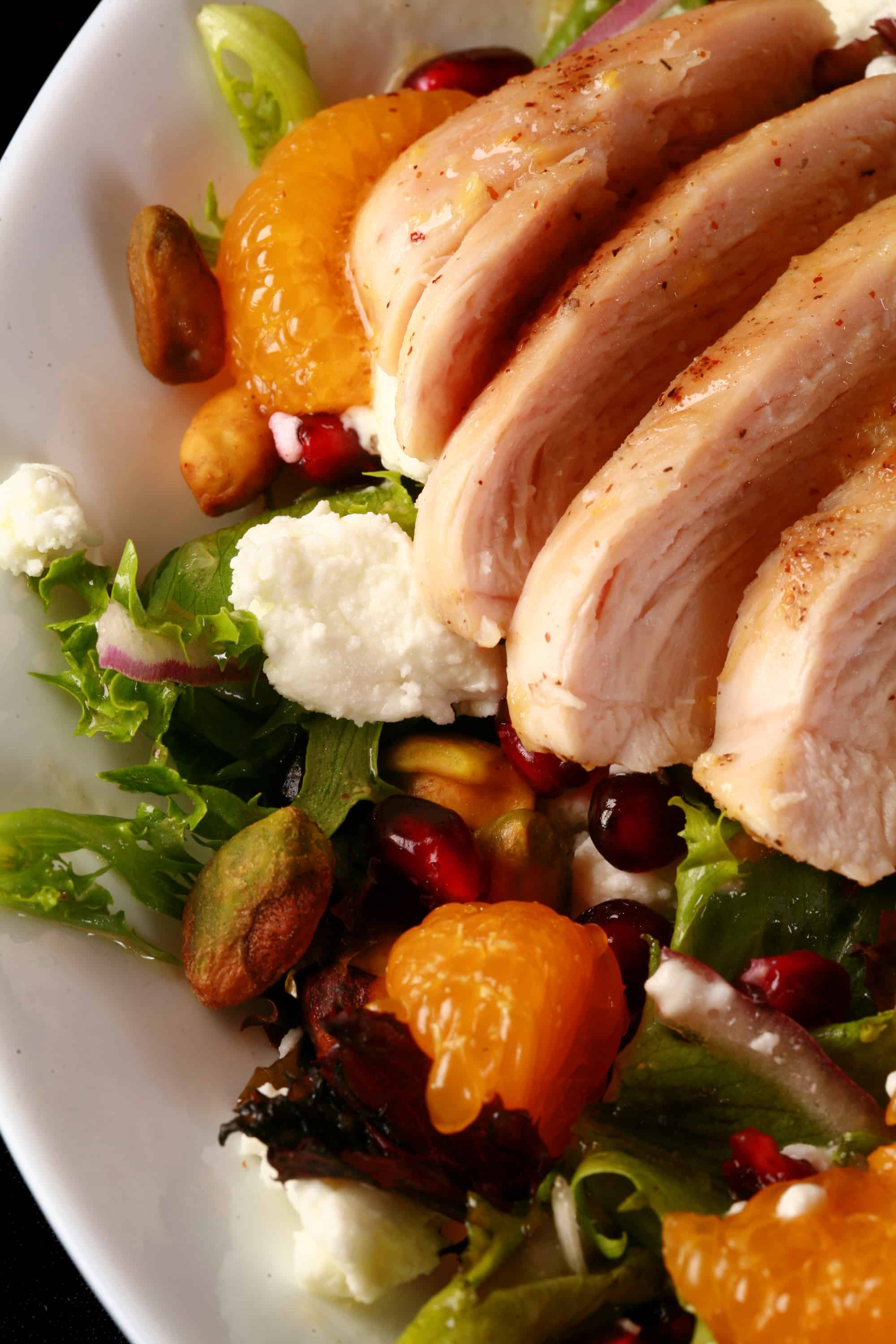 A bowl of Christmas salad with orange glazed chicken, mandarin orange segments, pomegranates, goat cheese, and pistachios.